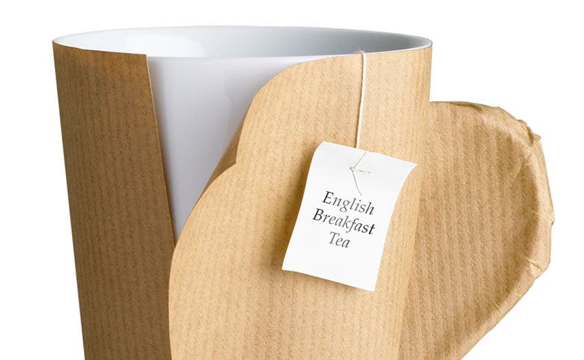 The image depicts the exterior of a residential property with a paved driveway on which a white van is parked, partially visible on the right side. The van is a standard commercial style vehicle operated by Hire Man and Van, designed for local rubbish removal services, with its side door closed. In the foreground, there is a neatly stacked pile of assorted waste materials, including black refuse sacks, flattened cardboard boxes, and a few loose items such as broken furniture parts and plastic containers. The waste appears to be prepared for collection, placed directly on the driveway surface, which is composed of grey concrete slabs. To the left, a section of the house's brick wall and a front garden with greenery are visible, providing a typical East Harling residential setting. The background shows a quiet street with parked cars and trees, characteristic of suburban neighbourhoods. The lighting is overcast, with soft, diffused daylight illuminating the scene evenly. The overall image captures a straightforward rubbish removal situation, with Hire Man and Van ready to assist in clearing household waste from this local property situated within the NR16 postcode area.